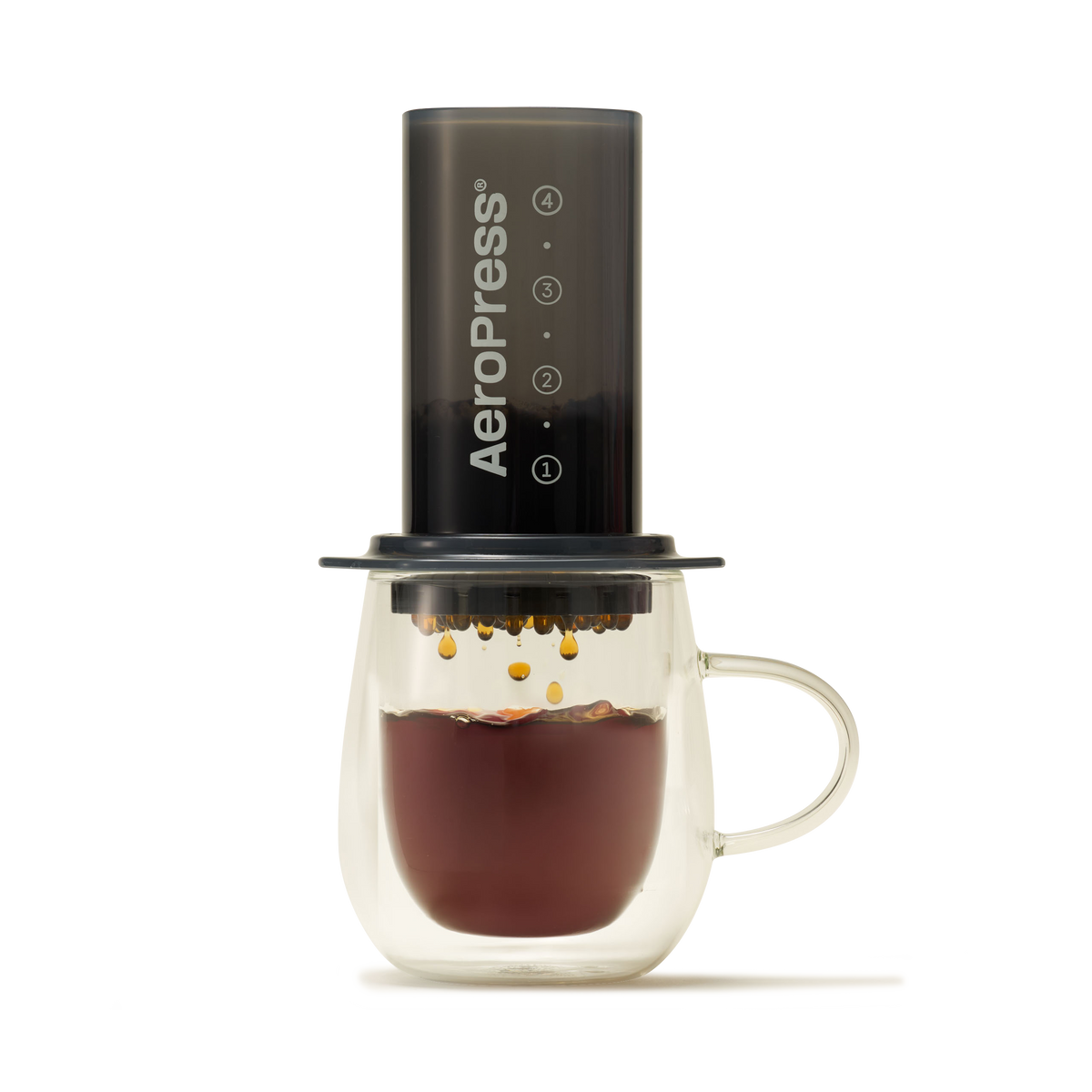 An AeroPress Coffee Maker is positioned over a transparent, double-walled glass mug, with freshly brewed, low-acidity coffee dripping from the brewer into the mug below. A compostable paper filter provides a clean and smooth brew, all arranged against a simple light gray background.