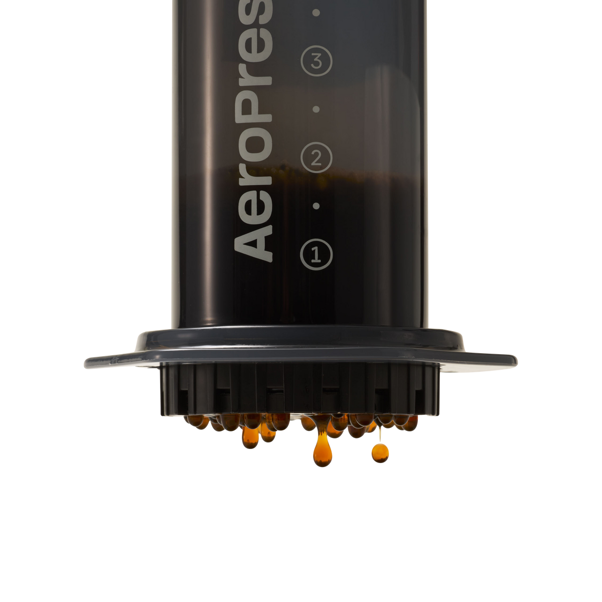 A translucent black AeroPress Coffee Maker is displayed against a white background, filled with dark coffee. Equipped with a compostable paper filter, the bottom filter cap releases freshly brewed coffee in small amber droplets, ensuring an excellent cup of coffee with low acidity.