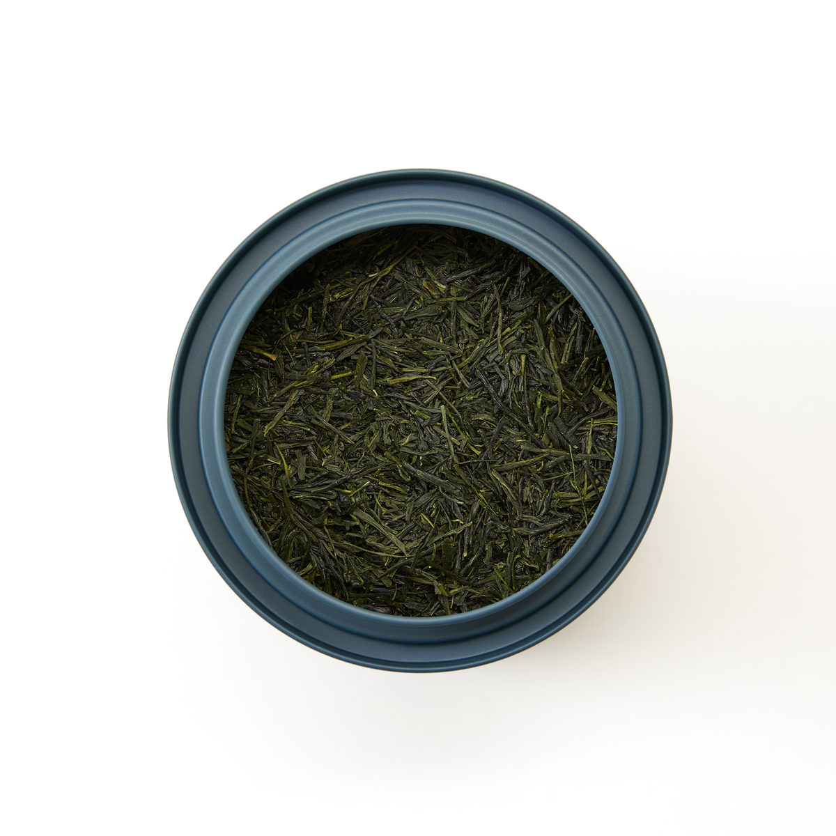 A round, dark container filled with Gyokuro Loose Leaf Tea is viewed from above against a plain white background. The vibrant green leaves hint at the rich umami flavor characteristic of this premium Japanese tea.