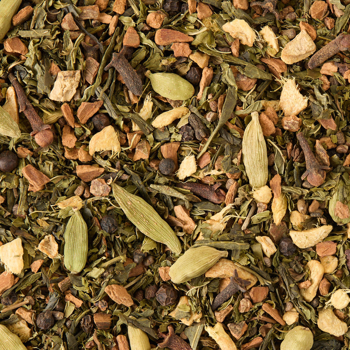 A close-up of Shanghai Chai Loose Leaf Tea reveals dried green leaves, cardamom pods, cloves, black peppercorns, and pieces of ginger and cinnamon, creating a textured and colorful blend—a perfect representation of spiced green tea.