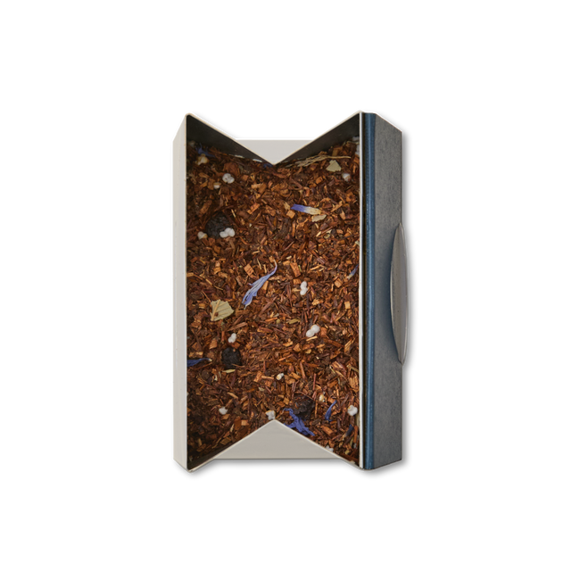 A rectangular, open-top box displays Blueberry Rooibos Loose Leaf Tea, a caffeine-free herbal blend with rooibos, flower petals, small white pieces, and hints of blueberry; the tea is viewed from above against a white background.