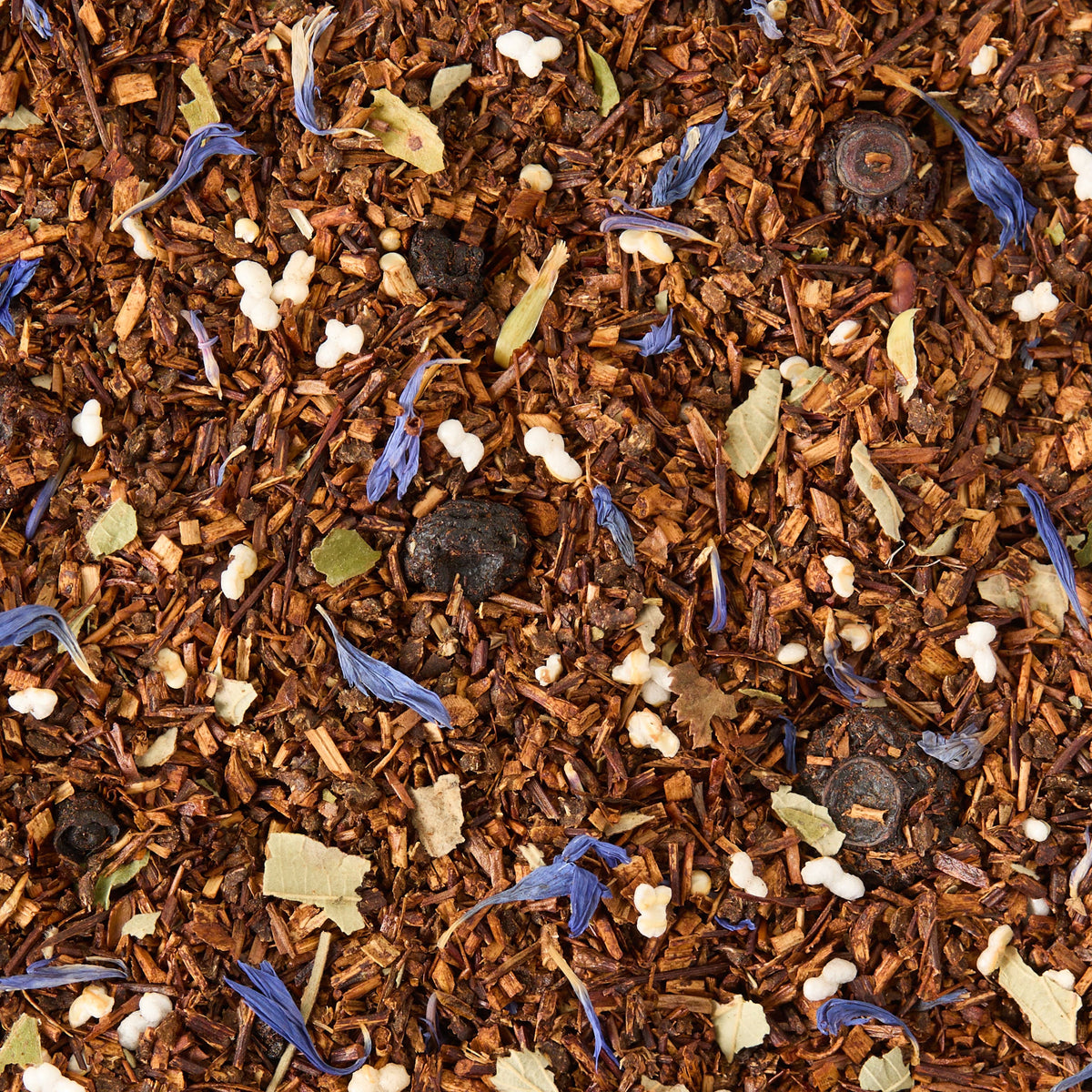 A close-up of Blueberry Rooibos Loose Leaf Tea, highlighting rooibos leaves, blue cornflower petals, white sugar pearls, dried berries, and small dried leaves—a naturally caffeine-free herbal infusion.