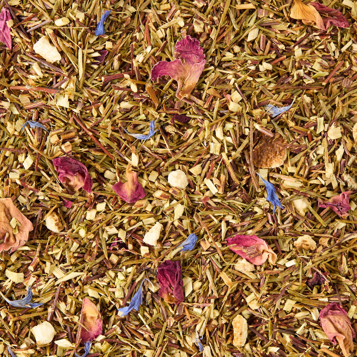 A close-up of Rainforest Rooibos Loose Leaf Tea shows a vibrant mix of loose herbal tea leaves with dried flower petals in shades of pink, purple, and blue, scattered among small greenish and brown plant pieces. This naturally caffeine free blend features green rooibos, making it perfect for any time of day.