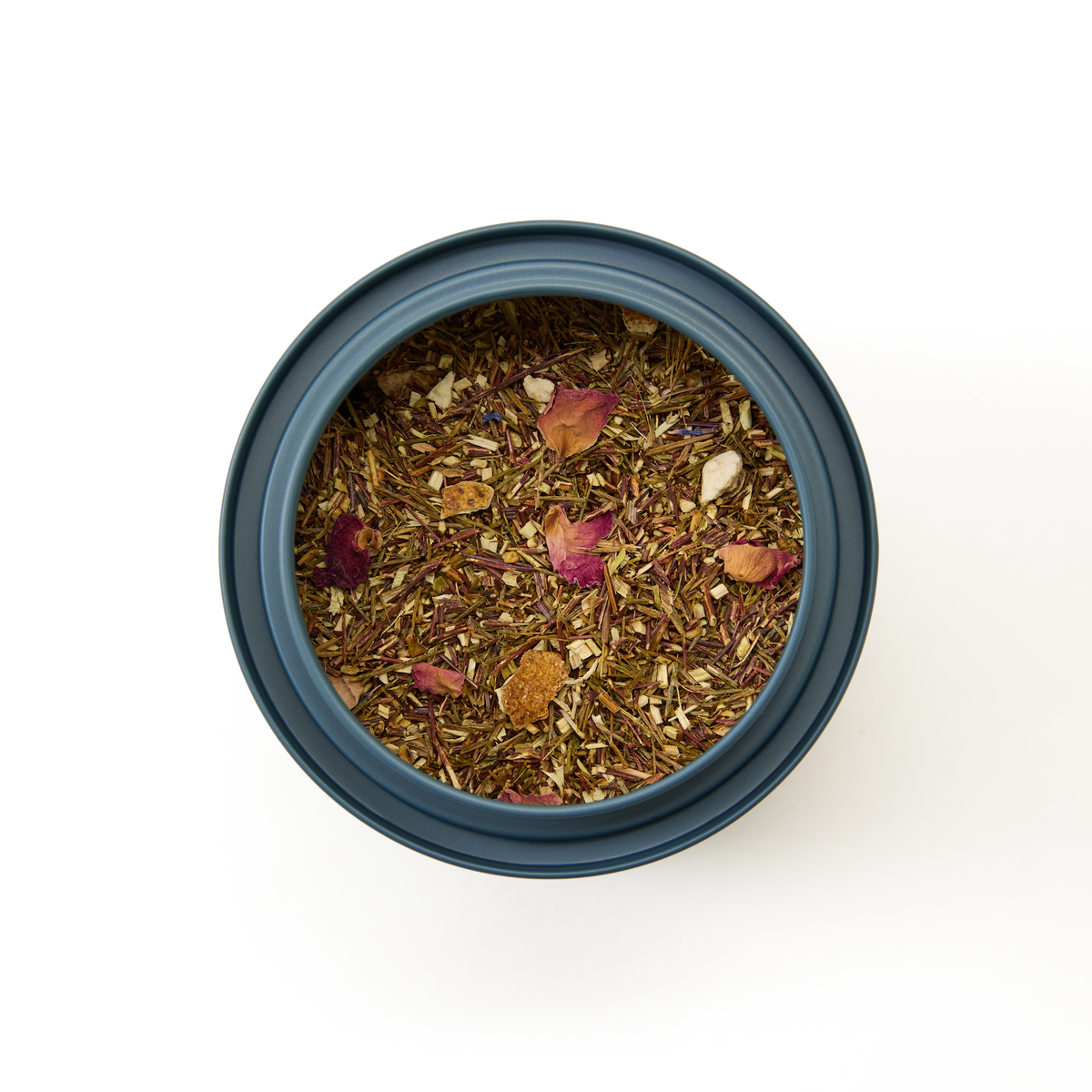 A round blue container viewed from above, filled with loose herbal tea leaves, dried petals, and small pieces of citrus peel against a white background—this vibrant blend is Rainforest Rooibos Loose Leaf Tea, a caffeine-free tea featuring green rooibos for a naturally uplifting tropical infusion.