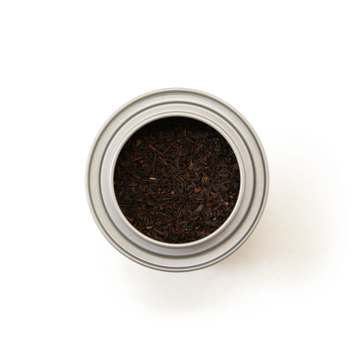 A top-down view of a round, silver tin labeled “English Breakfast Alice in Wonderland Mini Caddy,” filled with loose classic black tea leaves, on a white background.