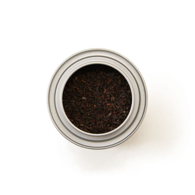 A top-down view of a round, silver tin labeled “English Breakfast Alice in Wonderland Mini Caddy,” filled with loose classic black tea leaves, on a white background.