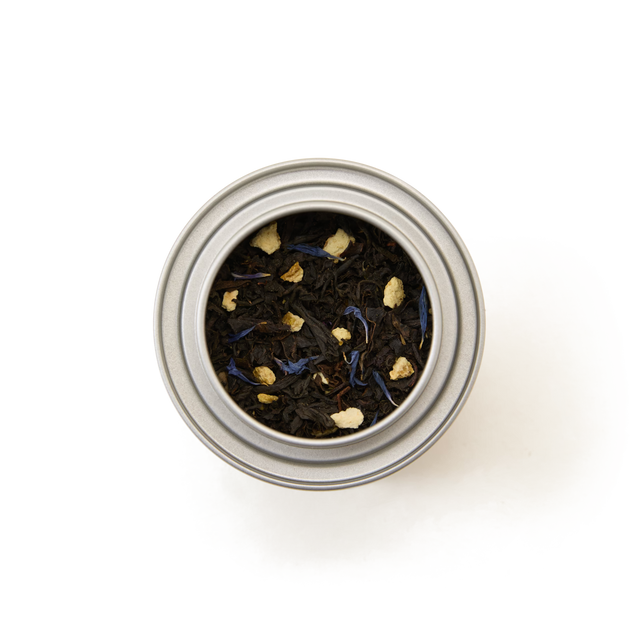 A top-down view of the Earl Grey Alice in Wonderland Mini Caddy, a metal tin filled with loose leaf black tea featuring dark leaves and visible pieces of dried fruit or spices, set against a white background—perfect for an elegant afternoon tea.