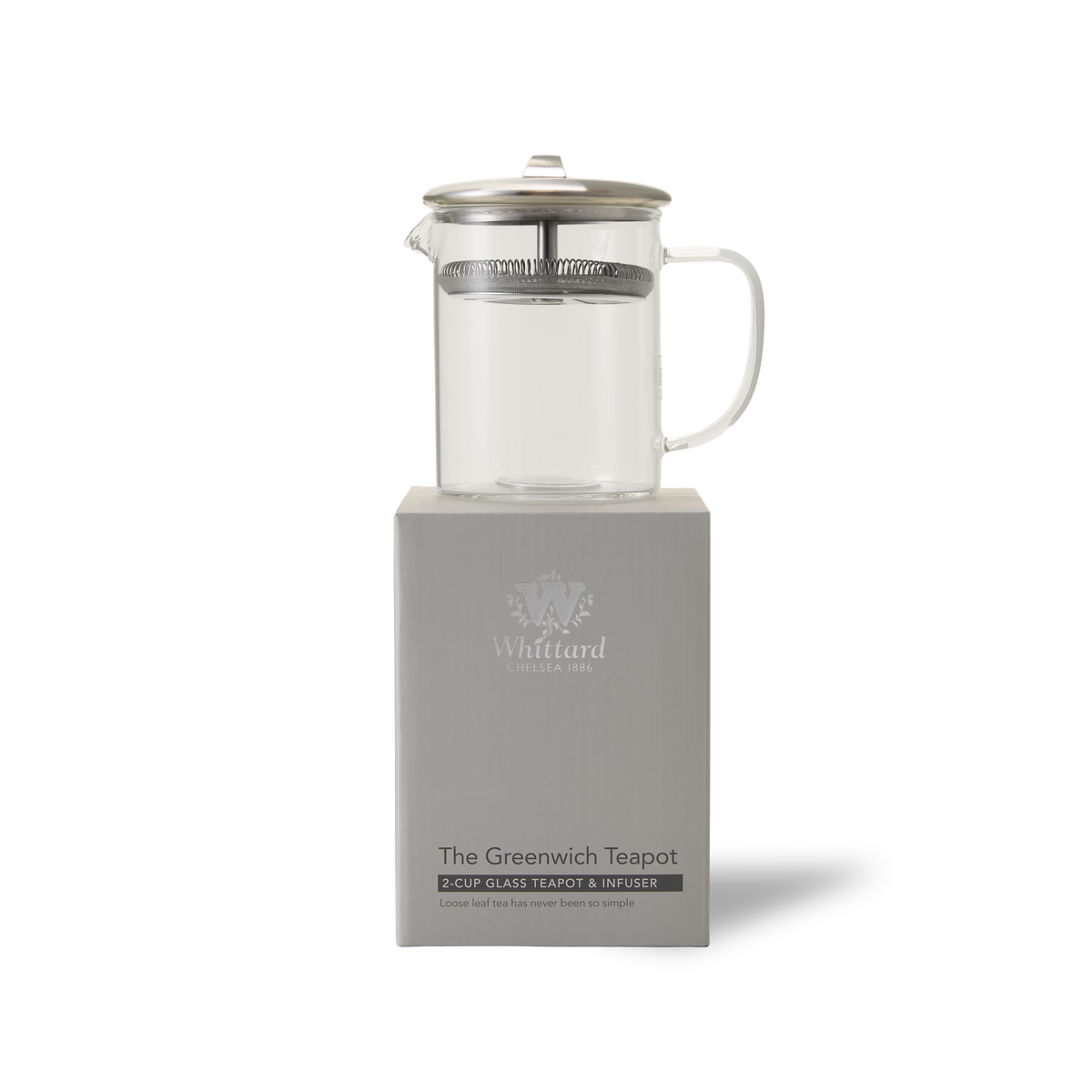 A clear glass teapot with a metal lid and infuser sits atop a gray box labeled The Greenwich Teapot by Whittard. The background is white and minimalistic.