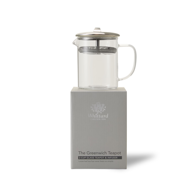 A clear glass teapot with a metal lid and infuser sits atop a gray box labeled The Greenwich Teapot by Whittard. The background is white and minimalistic.