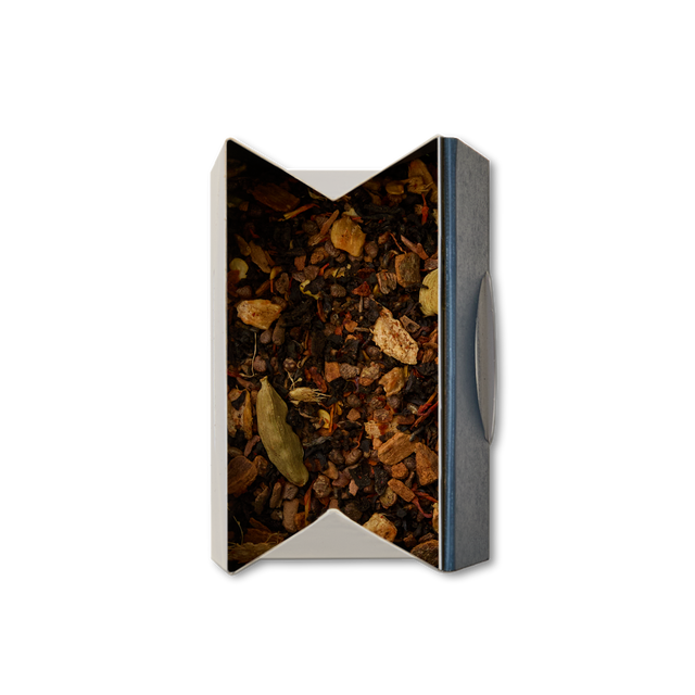 A top-down view of an open rectangular box containing Chilli Chai Loose Leaf Tea—a blend of dried tea leaves, spices, and herbs with visible cardamom pods and other ingredients—perfect for making a flavorful loose chai.