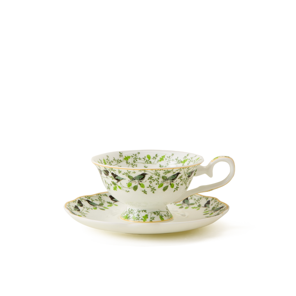 A delicate white teacup with a matching saucer, both decorated with green leaves and small black birds, accented by gold trim, set against a plain white background.