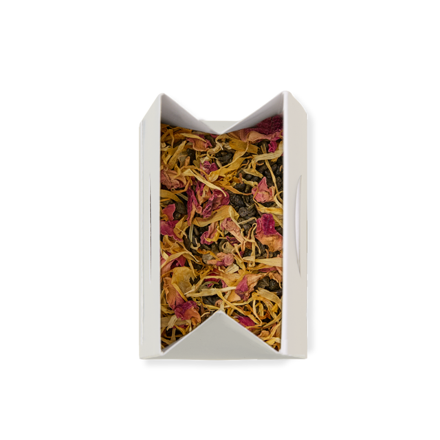 A white rectangular box filled with Passionfruit & Mango Loose Leaf Tea, showcasing a vibrant blend of dried tea leaves and colorful flower petals—yellow and pink—viewed from above against a plain white background. This inviting display highlights the premium loose leaf green tea, perfect for tea lovers.