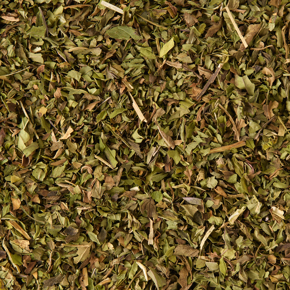 A close-up view of dried, chopped Peppermint Loose Infusion spread out evenly, revealing a mix of small, jagged leaves and slender stems.