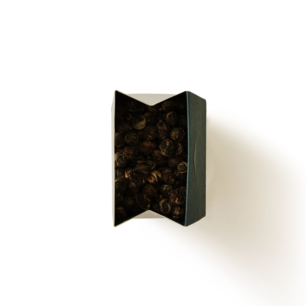 A top view of a dark, rectangular box partially open, revealing dried, round, brown objects inside—Jasmine Dragon Pearls Loose Leaf Tea—set against a plain white background with a shadow to the right.