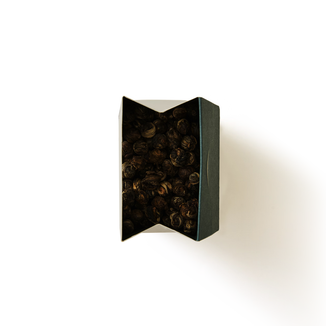 A top view of a dark, rectangular box partially open, revealing dried, round, brown objects inside—Jasmine Dragon Pearls Loose Leaf Tea—set against a plain white background with a shadow to the right.