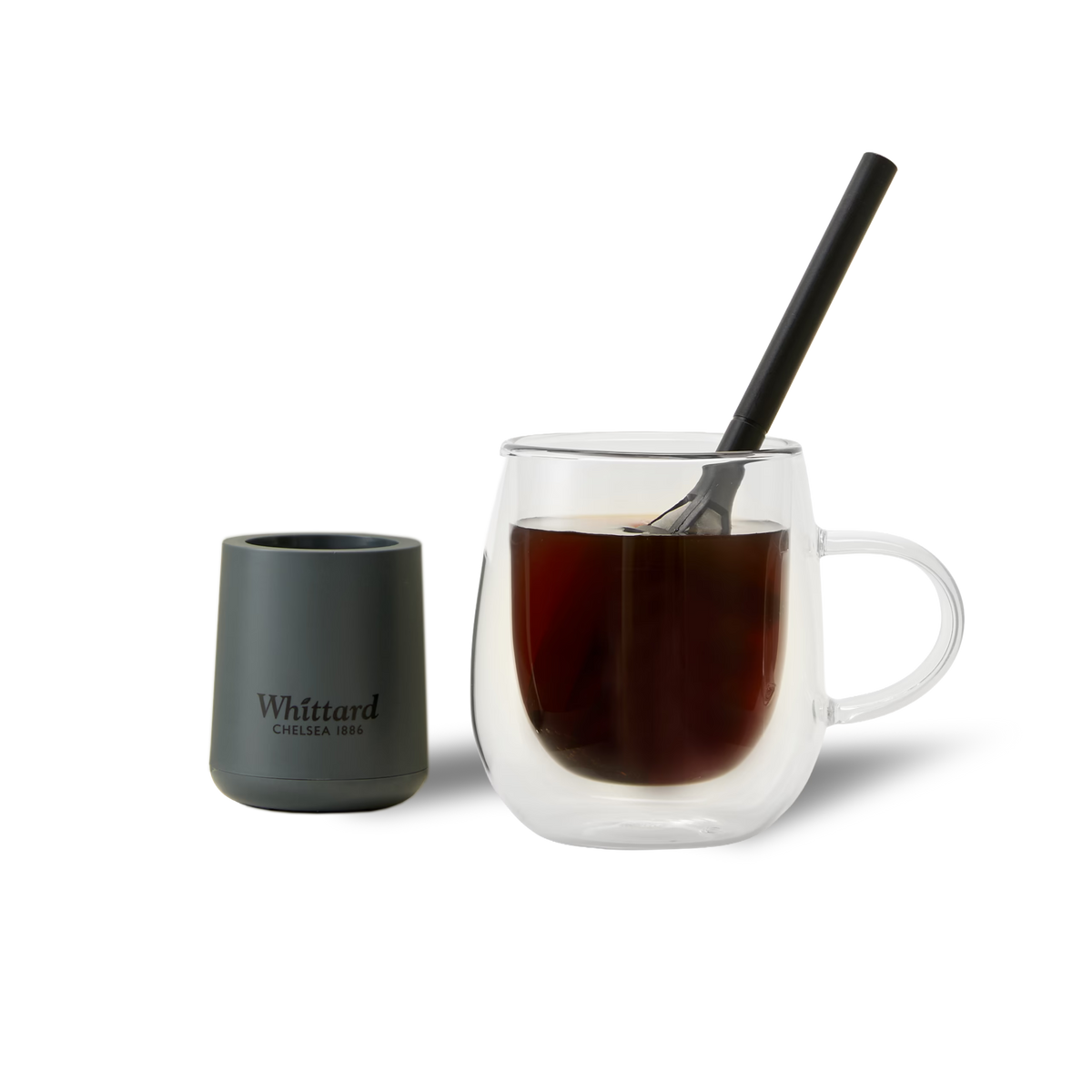 A glass mug filled with dark coffee and a black spoon, next to a matte black Whittard Chelsea 1886 container, on a white background.