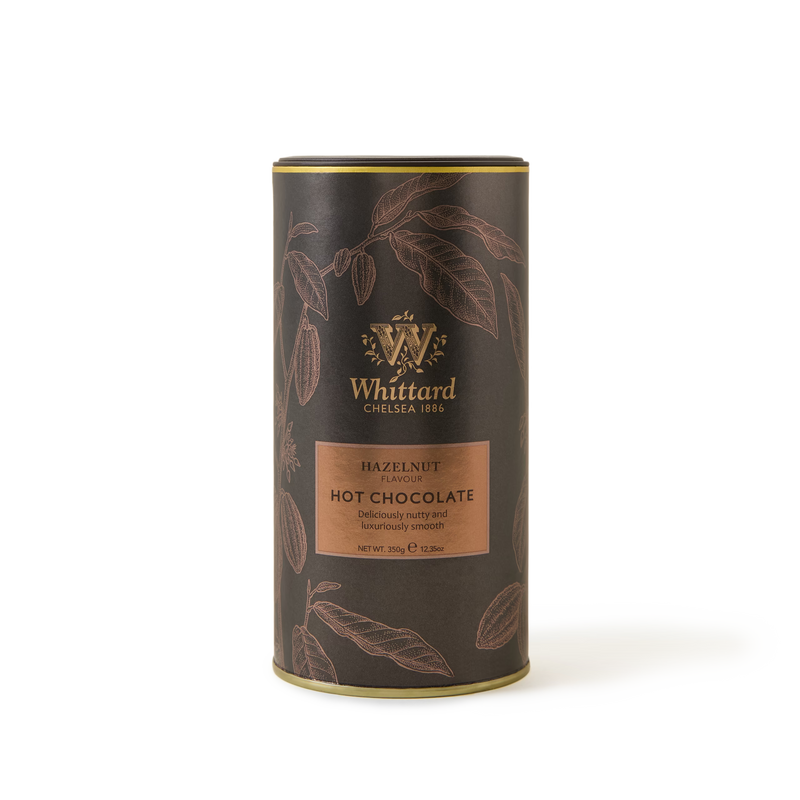 A cylindrical tin of Whittard Hazelnut Flavour Hot Chocolate. The brown tin features gold accents and botanical illustrations, with a label noting it is deliciously nutty and luxuriously smooth.