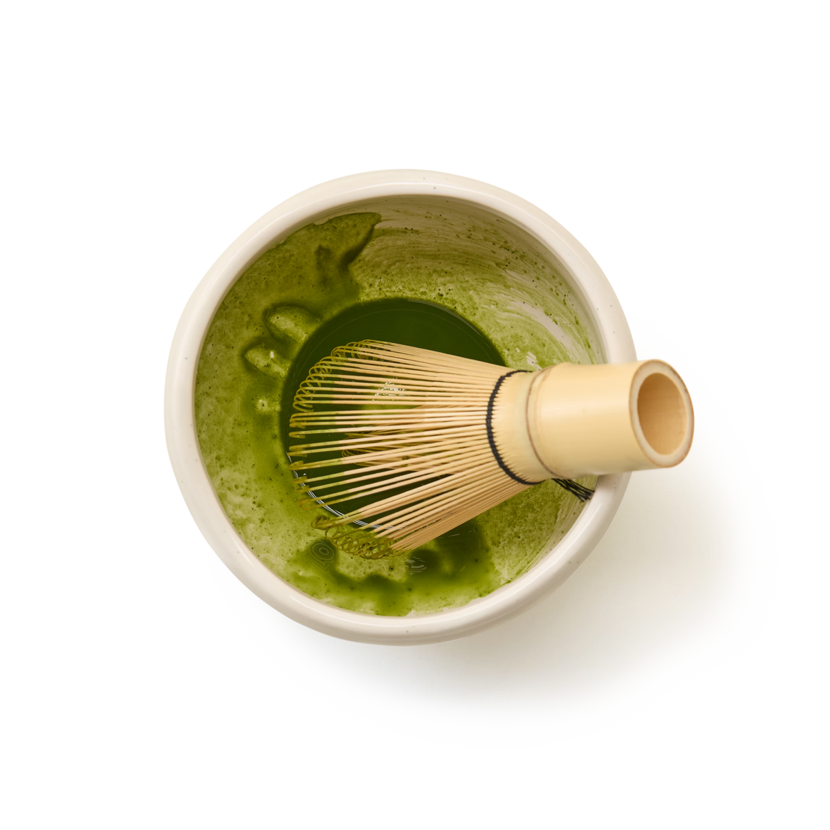 A bamboo whisk rests in a bowl of whisked matcha tea, seen from above on a white background.