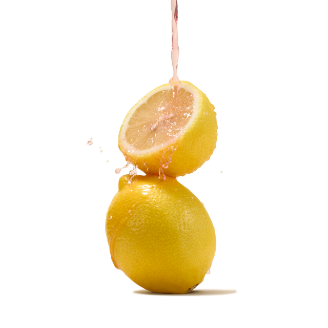 A stream of pink liquid splashes onto half a lemon balanced atop a whole lemon against a white background, capturing the lively essence of Pink Lemonade Flavour Instant Tea—a refreshing classic summer beverage.