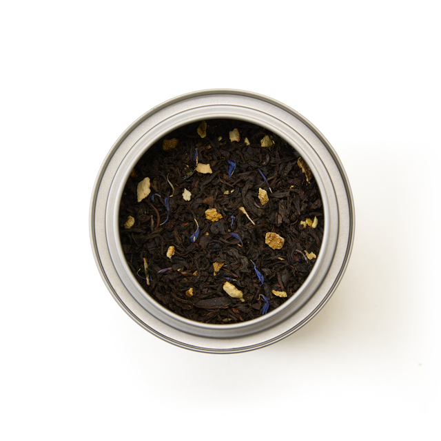 A top-down view of the Tea Discoveries Earl Grey Caddy, a round metal tin containing loose leaf Earl Grey tea made with dark Ceylon black tea leaves, delicate blue flower petals, subtle hints of bergamot, and small pieces of dried fruit or spice, set against a white background.