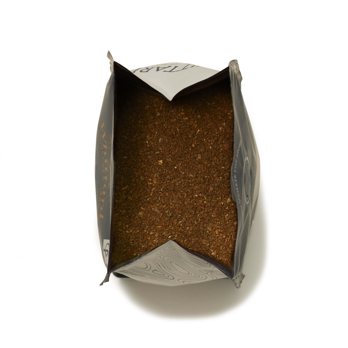 An opened bag of loose tea leaves, viewed from above with brown tea leaves filling most of the bag, sits alongside large ground coffee resembling the Guatemala Elephant Ground Coffee Valve Pack.