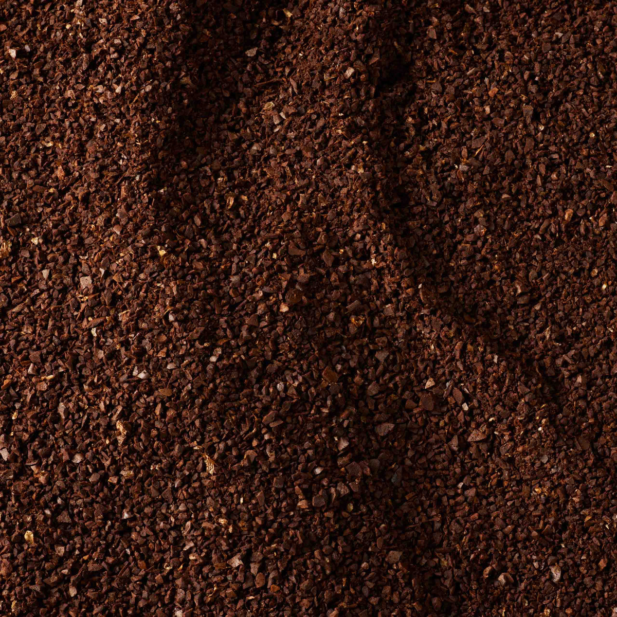 Close-up of a textured surface covered in dark brown coffee grounds, showing fine granules and an uneven distribution across the image. The rich color and texture evoke the robust flavors of Guatemala Elephant Ground Coffee Valve Pack, renowned for its large coffee beans grown near Lake Atitlán.