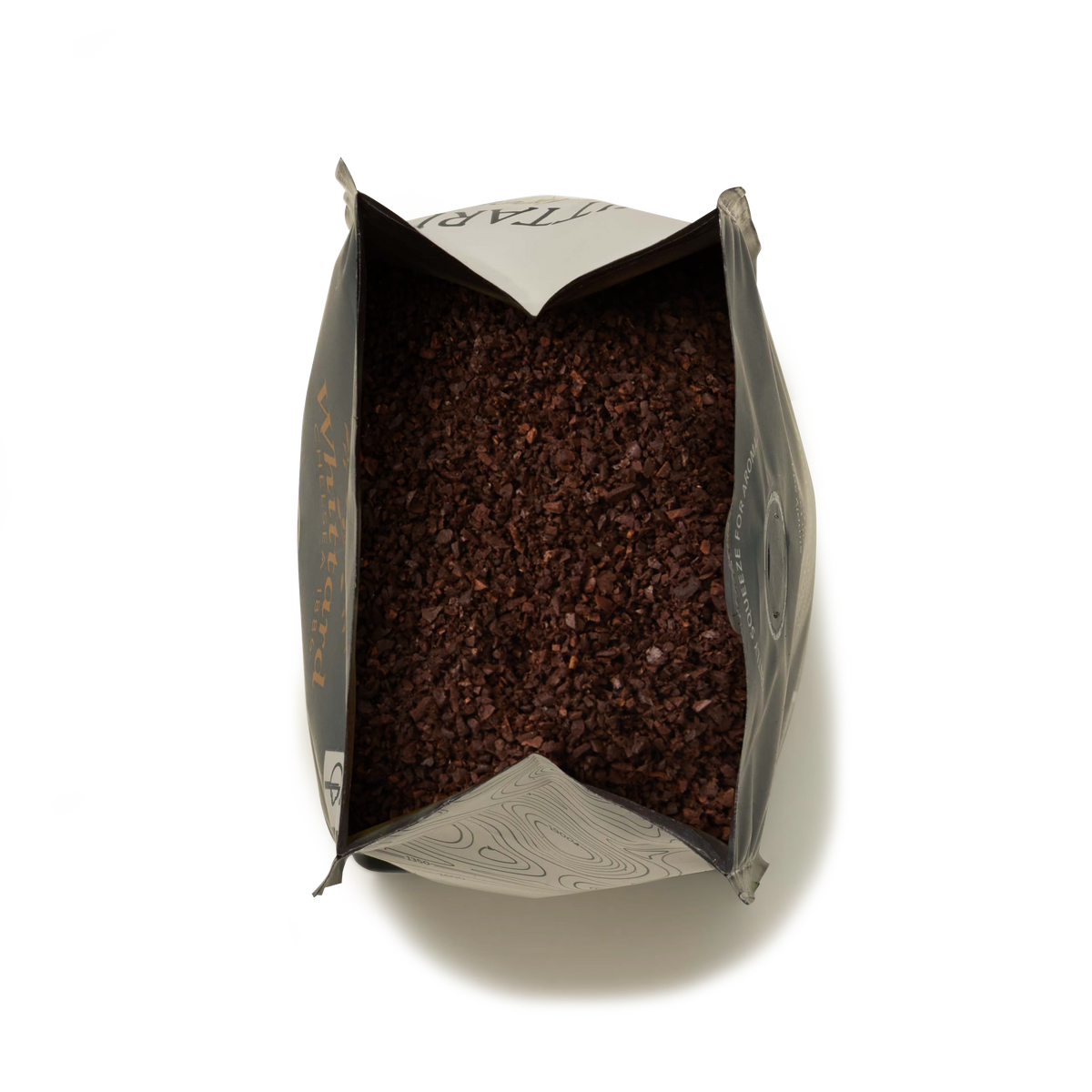 Open bag filled with dark brown, coarse Monsoon Malabar Ground Coffee, viewed from above, with the pack’s silver-gray sides partially folded outward against a white background—evoking the rich, aromatic texture characteristic of specialty coffees.