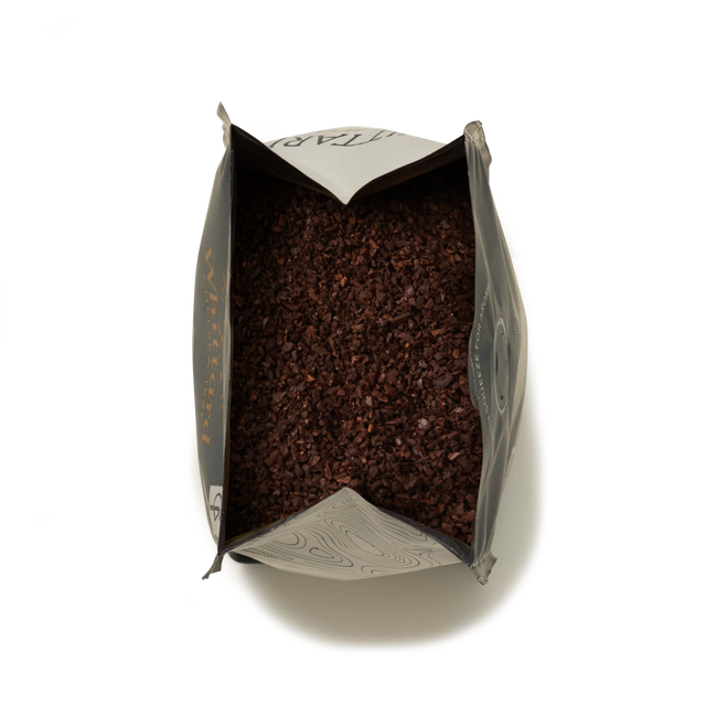 Open bag filled with dark brown, coarse Monsoon Malabar Ground Coffee, viewed from above, with the pack’s silver-gray sides partially folded outward against a white background—evoking the rich, aromatic texture characteristic of specialty coffees.