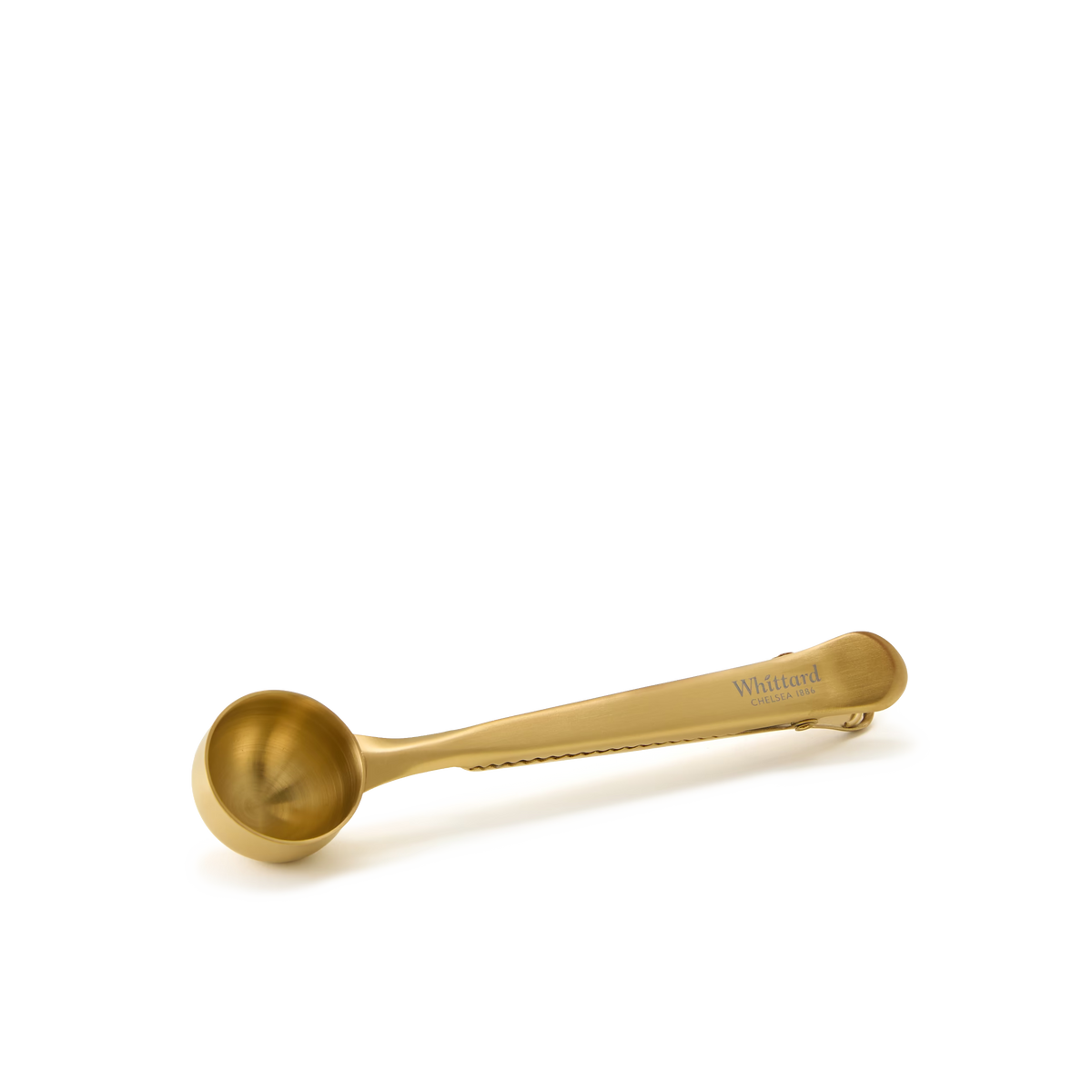 A gold-colored metal coffee scoop with a built-in bag clip, placed on a white background. The handle is engraved with Whittard..
