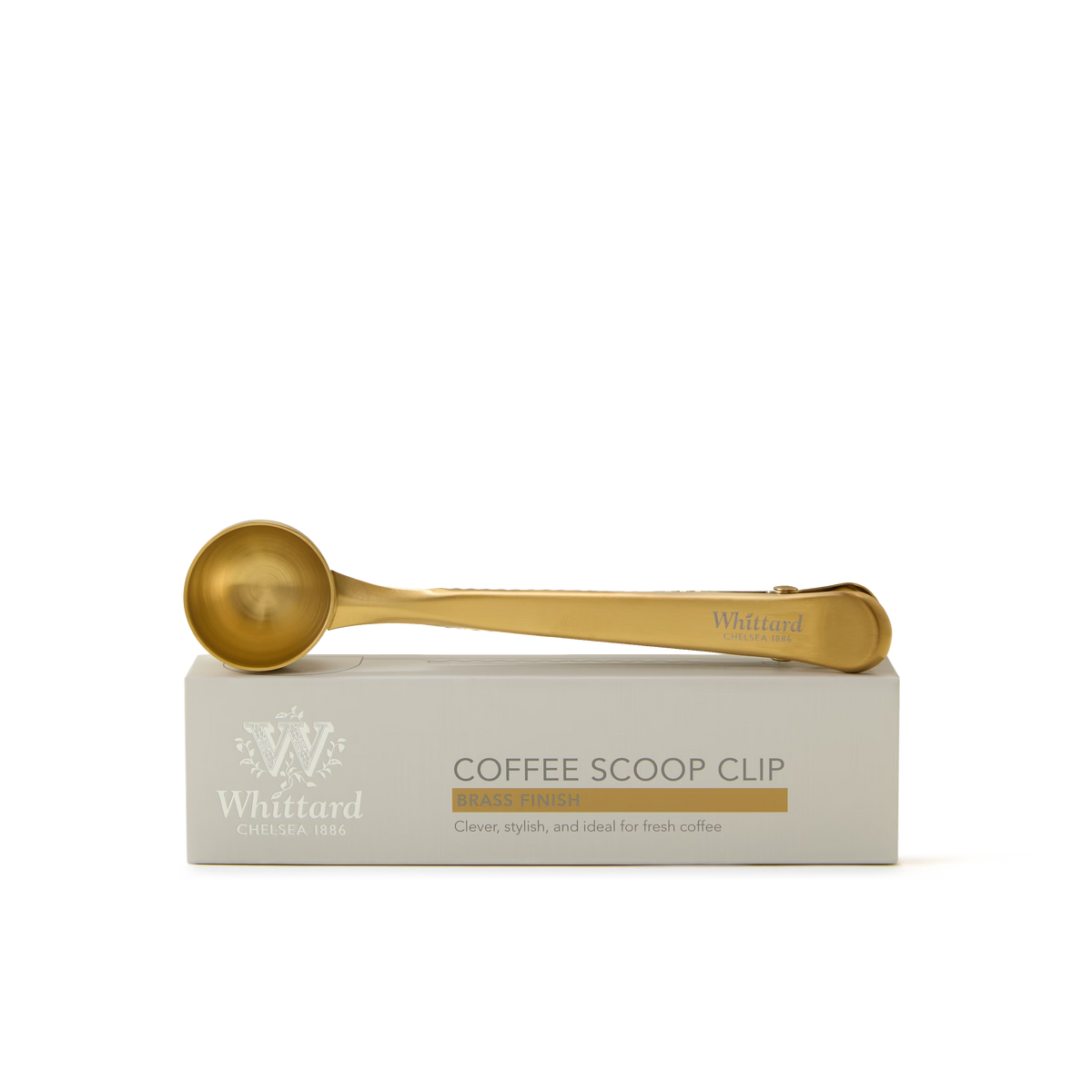 A gold-colored coffee scoop clip rests on a white Whittard box labeled Coffee Scoop Clip Brass Finish. The box features decorative text and logo detailing.