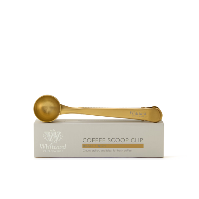 A gold-colored coffee scoop clip rests on a white Whittard box labeled Coffee Scoop Clip Brass Finish. The box features decorative text and logo detailing.