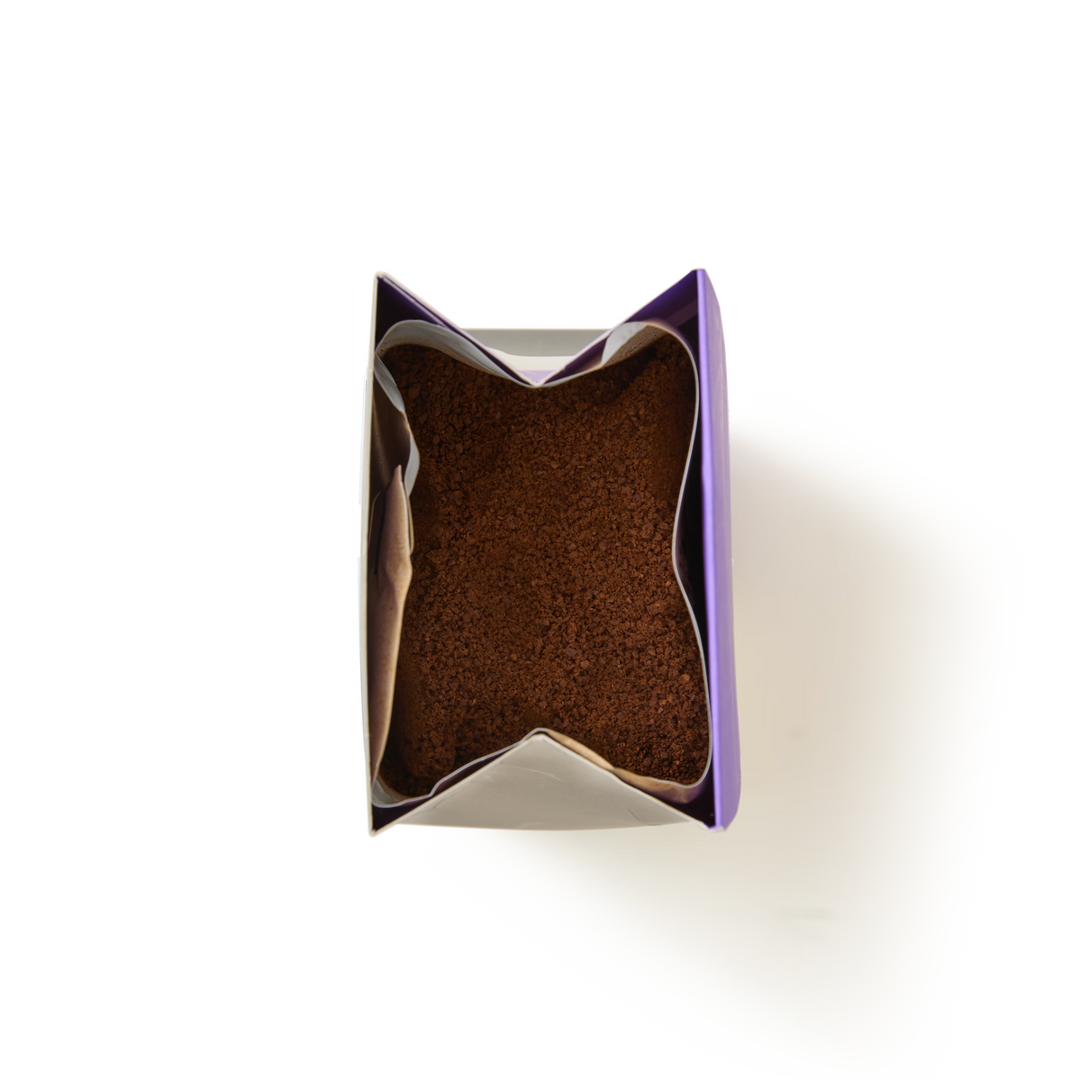 A top-down view of an open bag filled with brown, Chocolate Brownie Flavoured Ground Coffee, with the bag’s purple and silver edges visible against a white background.