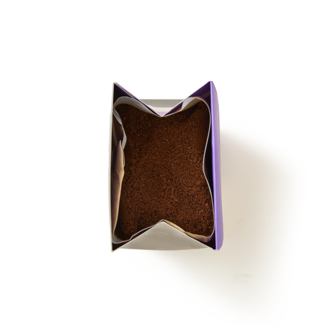 A top-down view of an open bag filled with brown, Chocolate Brownie Flavoured Ground Coffee, with the bag’s purple and silver edges visible against a white background.