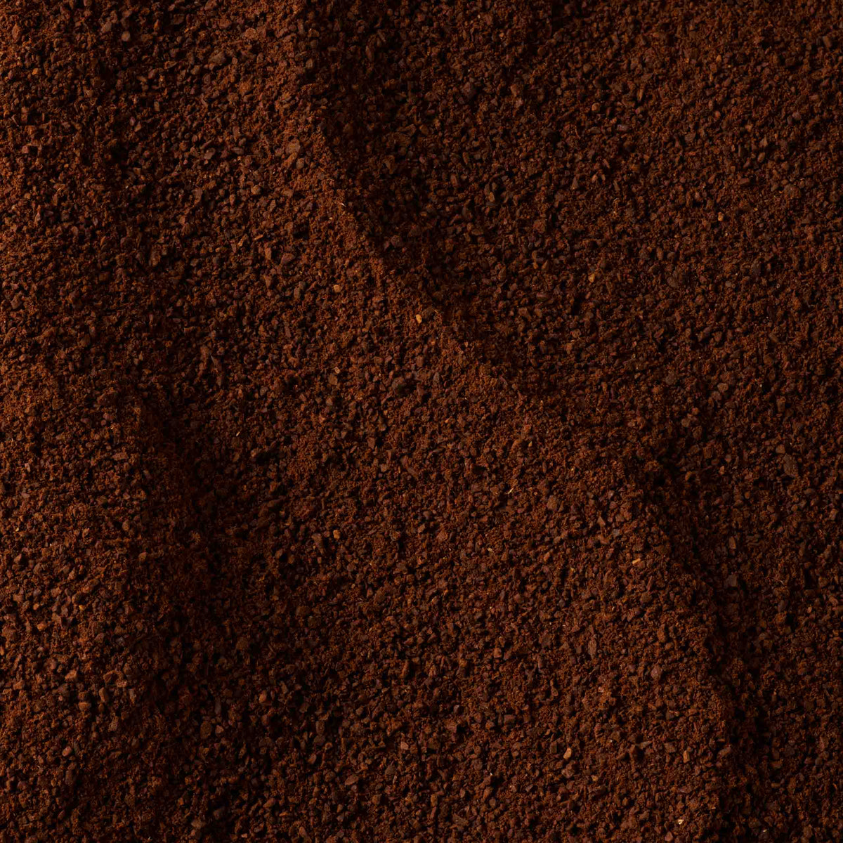 Close-up of Hazelnut Flavoured Ground Coffee with a dark brown, coarse texture reminiscent of a French café noisette. The surface appears slightly uneven, revealing small granules and subtle shadows.