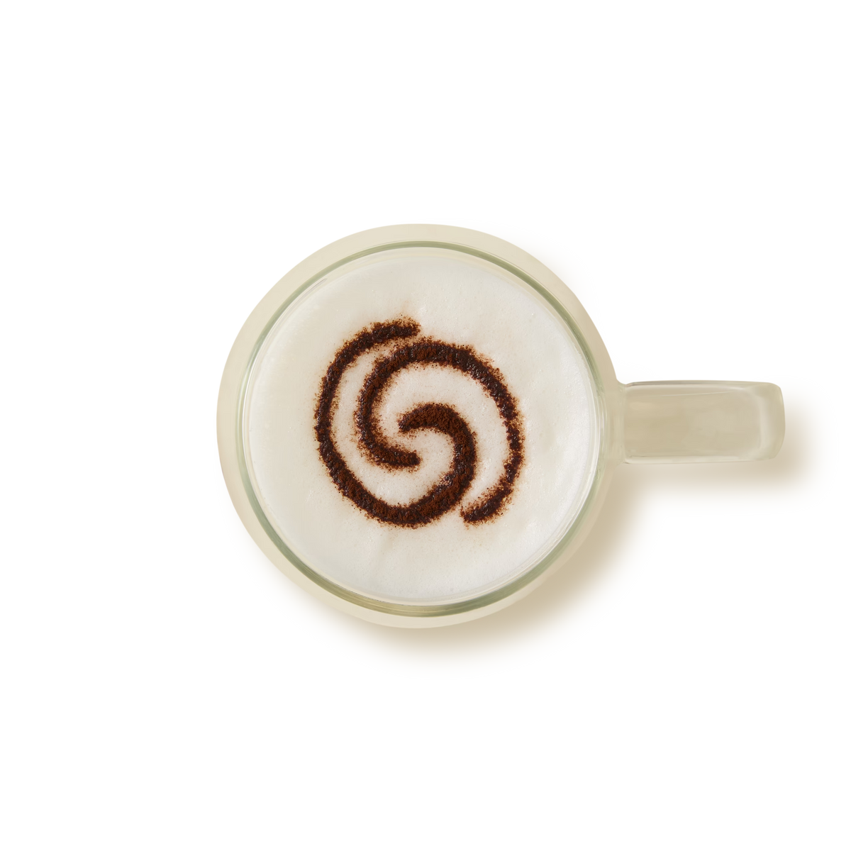 Top-down view of a glass coffee cup filled with frothy milk, featuring a swirling spiral pattern made with cocoa or cinnamon powder on the foam. The cup handle is on the right against a white background.