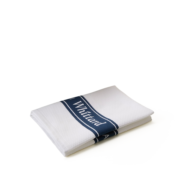 A neatly folded white towel with a textured pattern, featuring a navy blue stripe and the word White visible on the stripe, set against a plain white background.