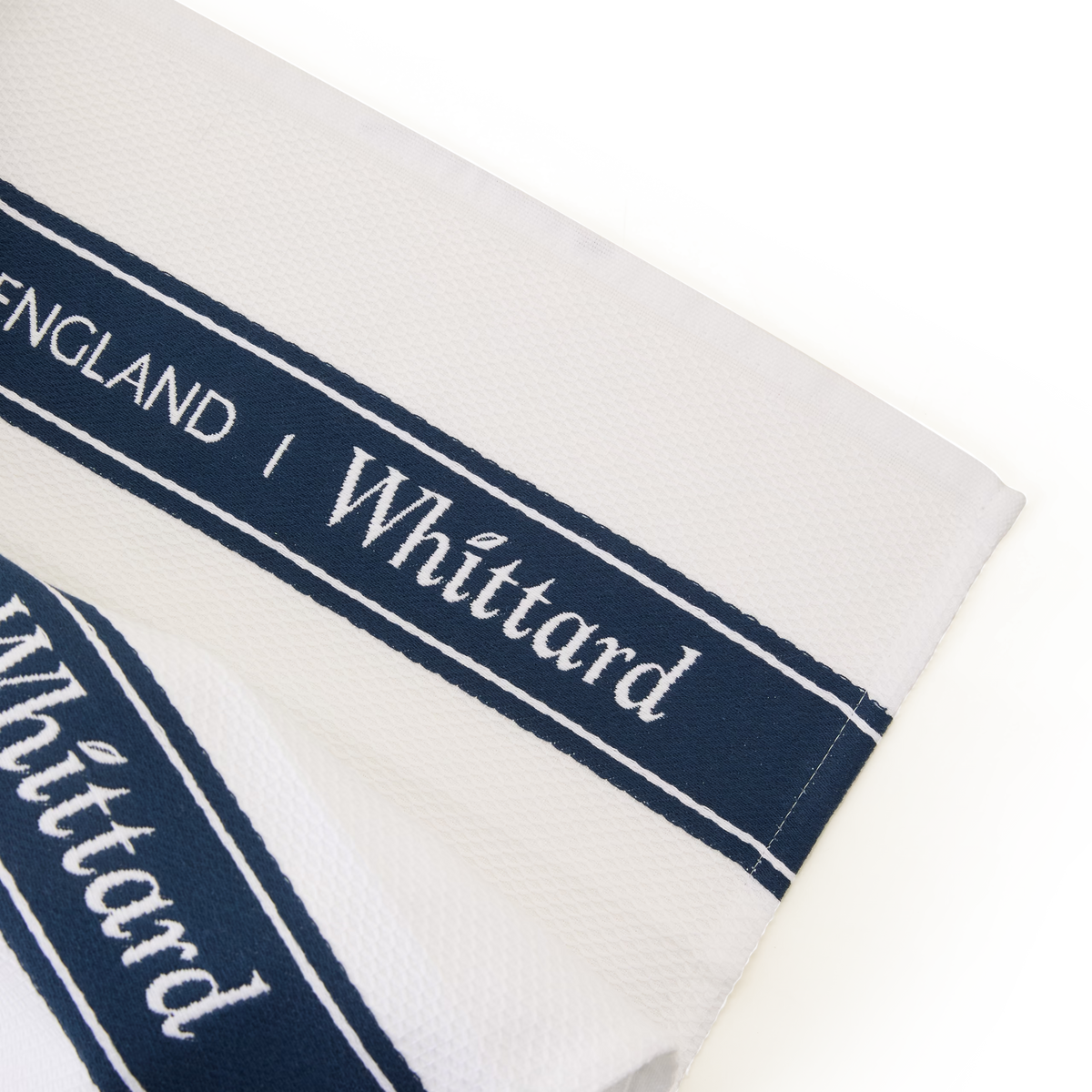 A close-up of a white textured cloth with navy blue bands that have the word Whittard and ENGLAND printed in white text.
