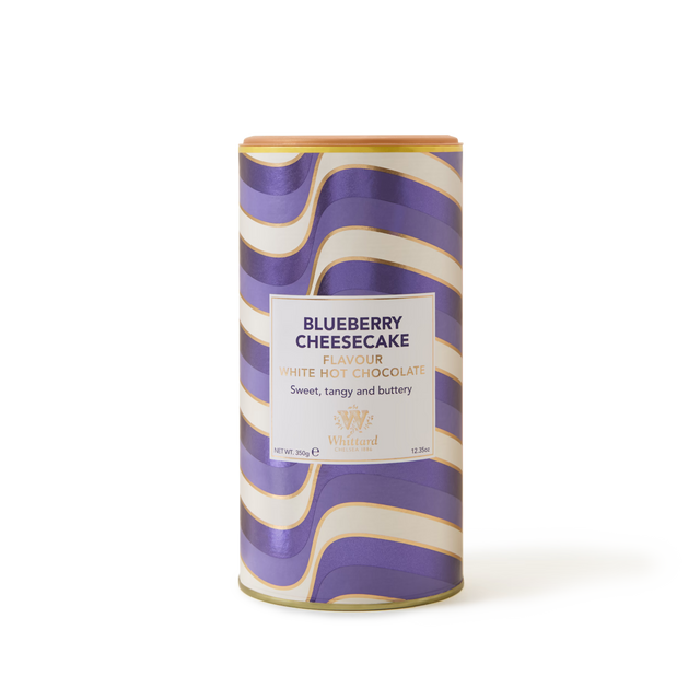A cylindrical tin of Whittard Blueberry Cheesecake flavored white hot chocolate, with a wavy purple, gold, and white design and text label on the front.
