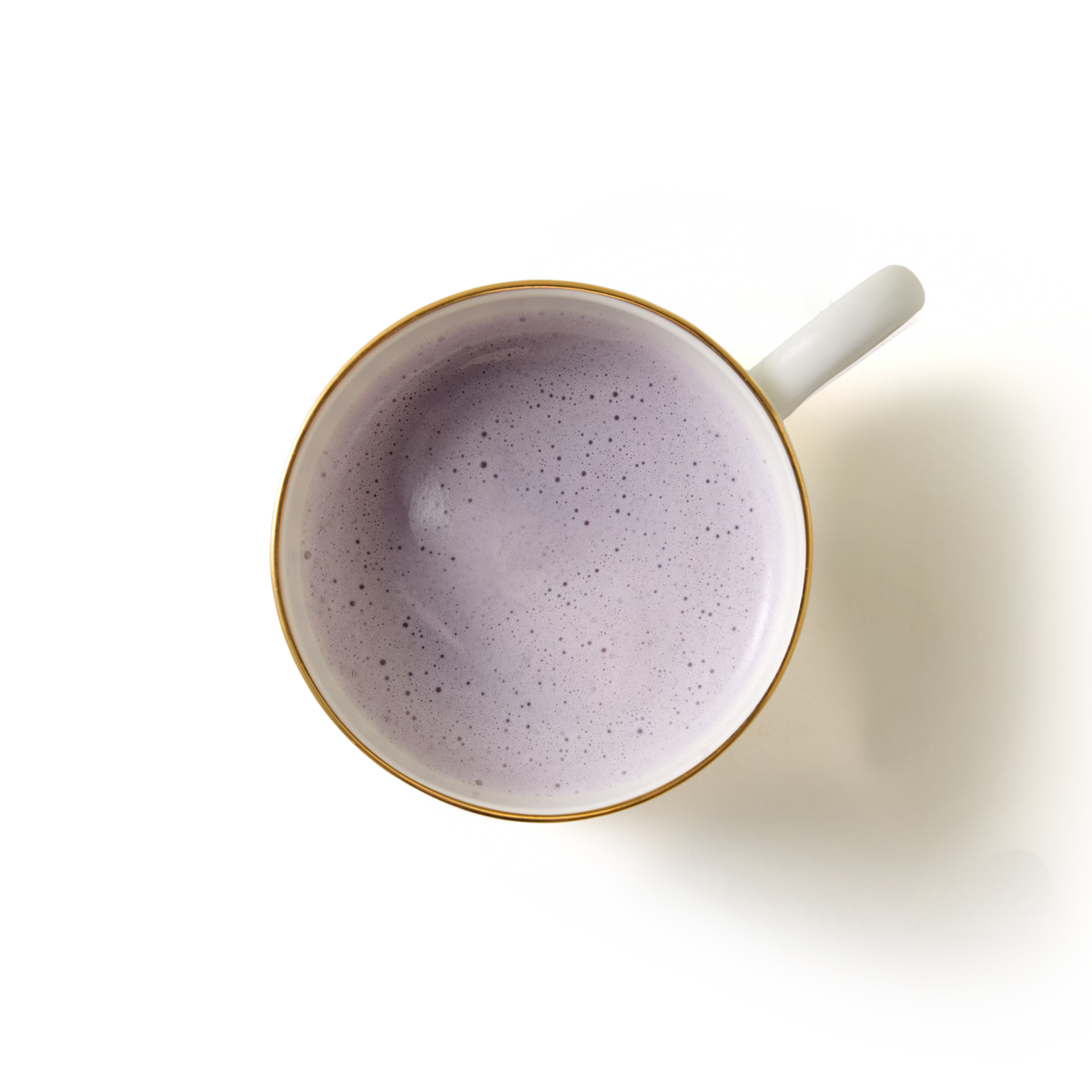 Top view of a cup filled with a creamy, light purple beverage, possibly a latte or smoothie, against a plain white background. The cup has a white handle and a gold rim.