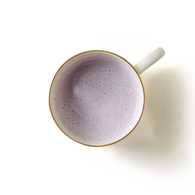 A white mug with a gold rim, filled with Blueberry Cheesecake Flavoured Hot Chocolate topped with light purple foam, viewed from above against a plain white background.