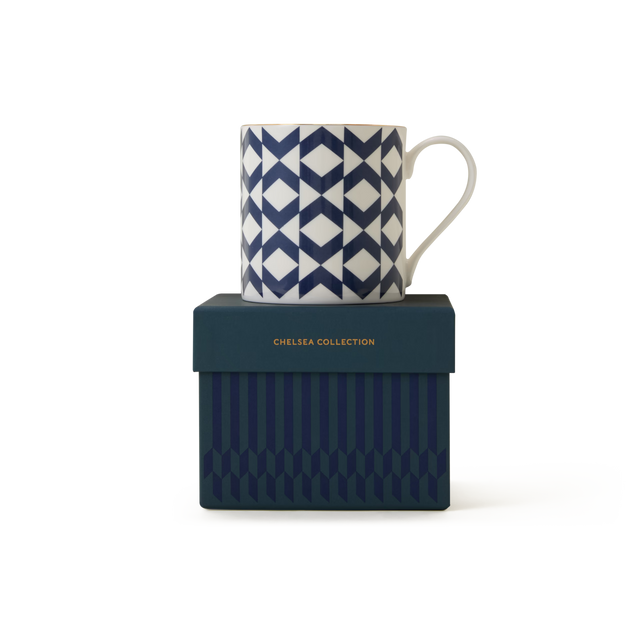 A white The Regency Mug featuring a blue geometric pattern and refined 22ct gold detailing rests atop a dark blue box embossed with “CHELSEA COLLECTION” in gold lettering. Both the mug and the box showcase coordinated pattern designs, embodying the enduring elegance of British ceramics.