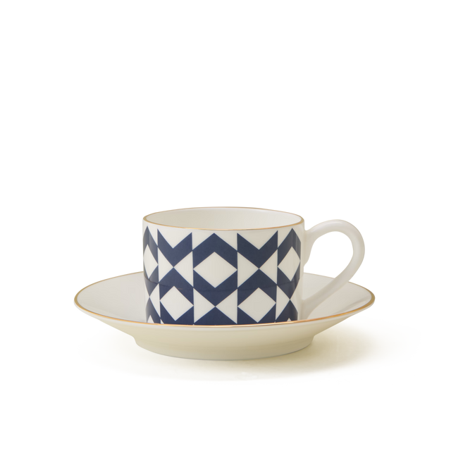 A white porcelain cup with a handle, featuring a navy blue geometric pattern, sits on a matching white saucer with a thin gold rim, against a plain white background.