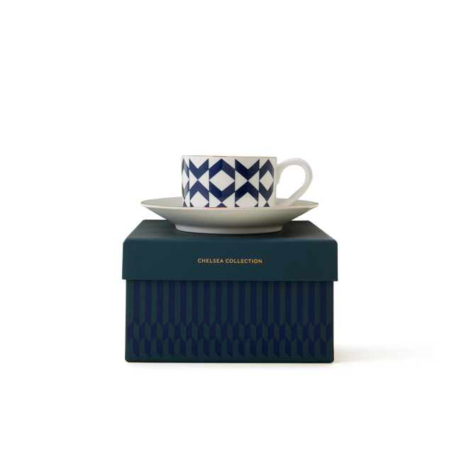 A white cup with a blue geometric pattern sits on a saucer, both placed on top of a dark blue box labeled Chelsea Collection in gold text. The background is plain white.