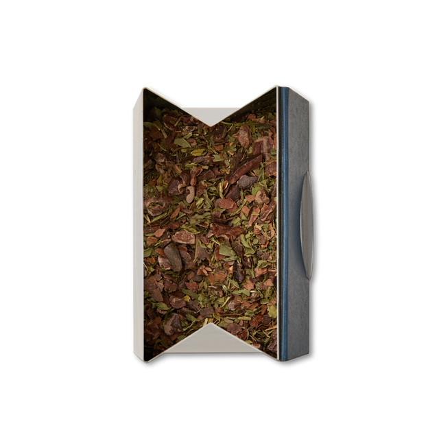 A rectangular box of Cacao & Mint Loose Infusion, filled with a blend of dried herbs and leaves seen from above. The open, angled top reveals the infusion's contents, framed by a simple dark rim on one side.