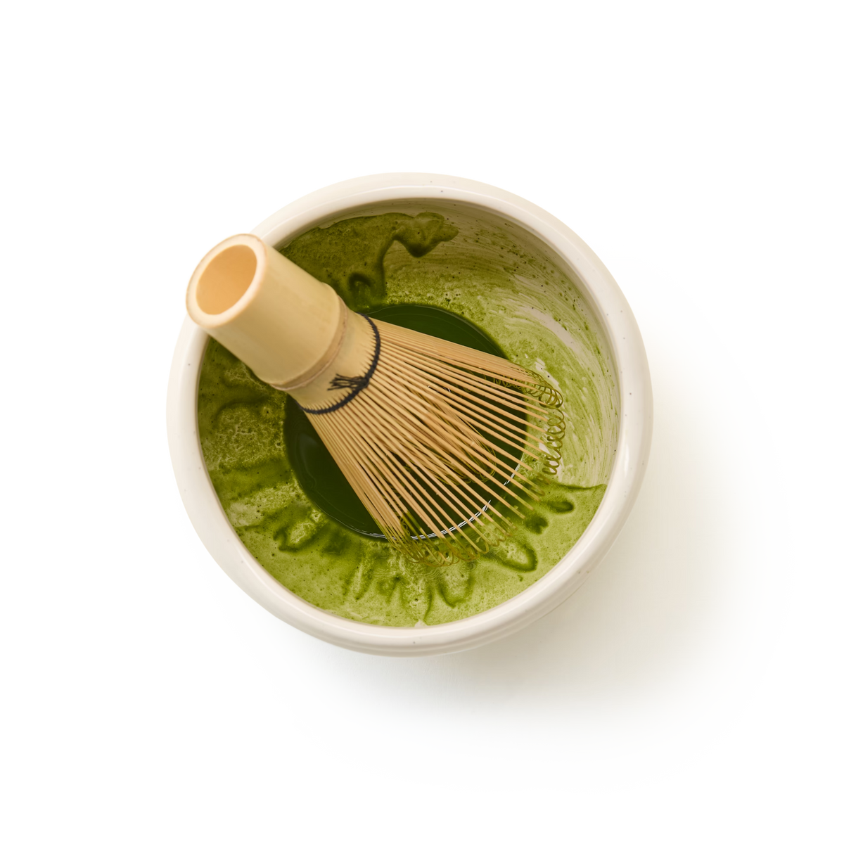 A Bamboo Matcha Whisk, an essential tool for traditional matcha preparation, sits in a bowl with vibrant green matcha tea, viewed from above on a white background.