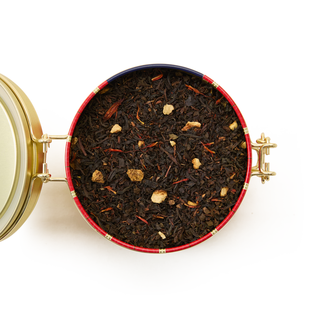 A round Christmas Tea Clip Top Tin with a gold hinged lid, filled with loose leaf black tea blended with small dried fruit and flower pieces, viewed from above on a white background.