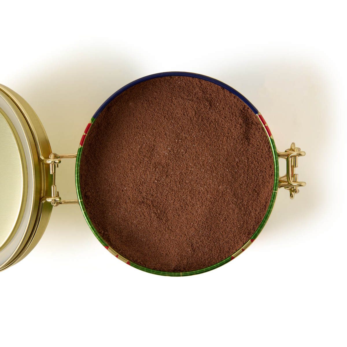 An open round tin filled with finely ground brown cocoa powder is shown from above against a plain white background. The lid is hinged to the left side.
