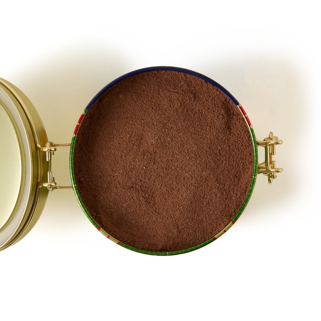A round, open tin of the Luxury Hot Chocolate Clip Top Tin, filled with fine brown powder and viewed from above. The tin features a gold lid hinged to the side and secured with a latch closure. Presented in elegant packaging that elevates any indulgent moment, it’s perfect for preparing creamy hot chocolate. Set against a plain white background.