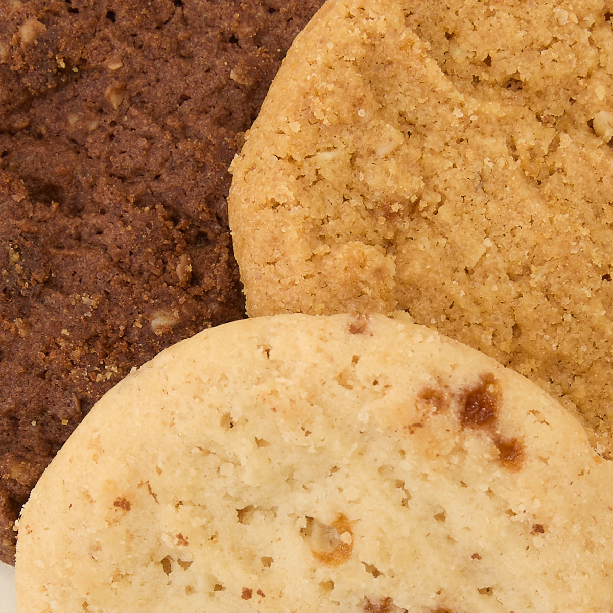 Three assorted cookies overlap each other. One is chocolate, one is oat or peanut butter, and one is a light-colored sugar or shortbread cookie. All have a crumbly texture and visible crumbs.