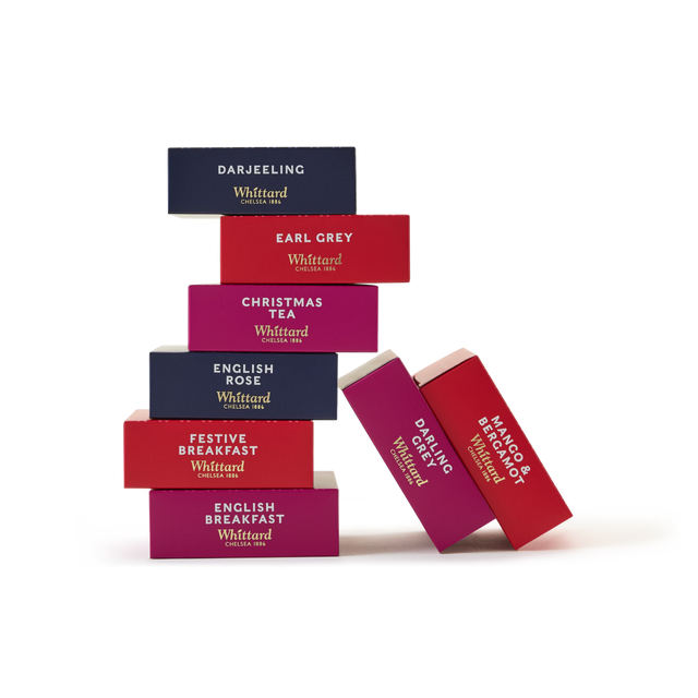 Seven vibrant tea boxes from A Feast of Tea, each marked with a unique tea flavor, are artfully stacked in a staggered display against a clean white background. An ideal Christmas tea gift for enthusiasts, this collection showcases boxes in blue, red, and pink adorned with refined gold and white lettering.